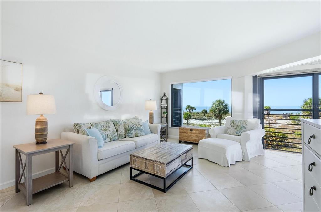 A bright living room features a sofa, armchair, coffee table, and large windows overlooking the beach — 5000 GASPARILLA ROAD UNIT BC205, BOCA GRANDE, FL