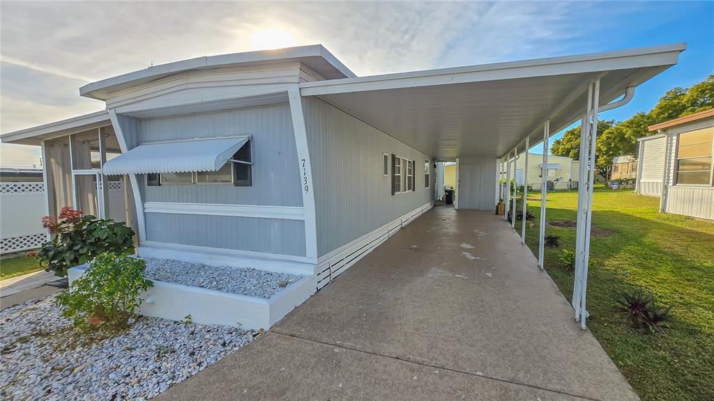 A mobile home exterior features a long concrete carport, light gray siding, and a gravel garden — 7139 EL CONQUISTADOR STREET, ZEPHYRHILLS, FL