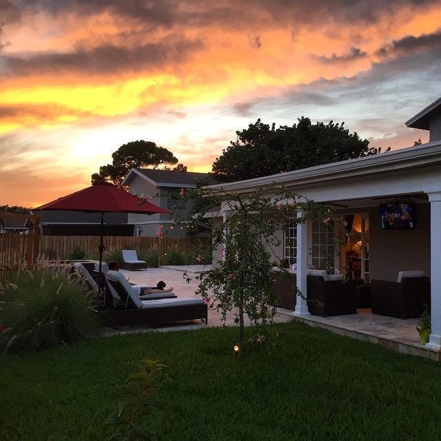 A backyard patio features lounge chairs, a red umbrella, and a covered porch at sunset — 4610 Pinetree Drive, Delray Beach FL 33445, DELRAY BEACH, FL