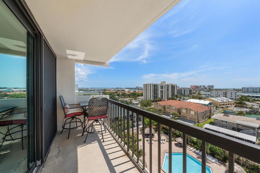 A balcony with metal railings overlooks a swimming pool, residential buildings, and a distant bay — 400 ISLAND WAY UNIT 904, CLEARWATER BEACH, FL