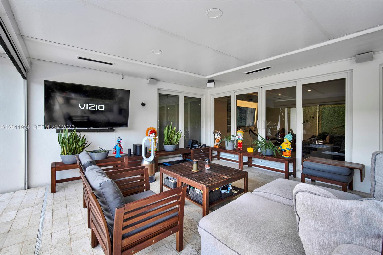 A covered outdoor patio features a wall-mounted television, wooden furniture, and several potted plants — 21015 NE 19th Ct, Miami FL 33179, MIAMI, FL
