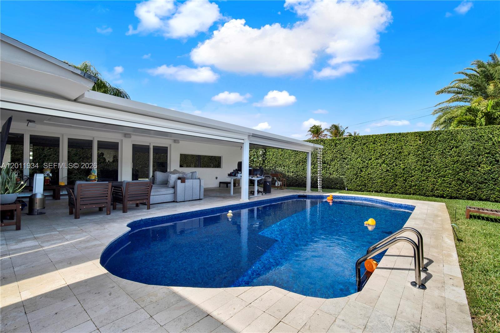 A backyard features a swimming pool, a covered patio with outdoor furniture, and a tall hedge — 21015 NE 19th Ct, Miami FL 33179, MIAMI, FL