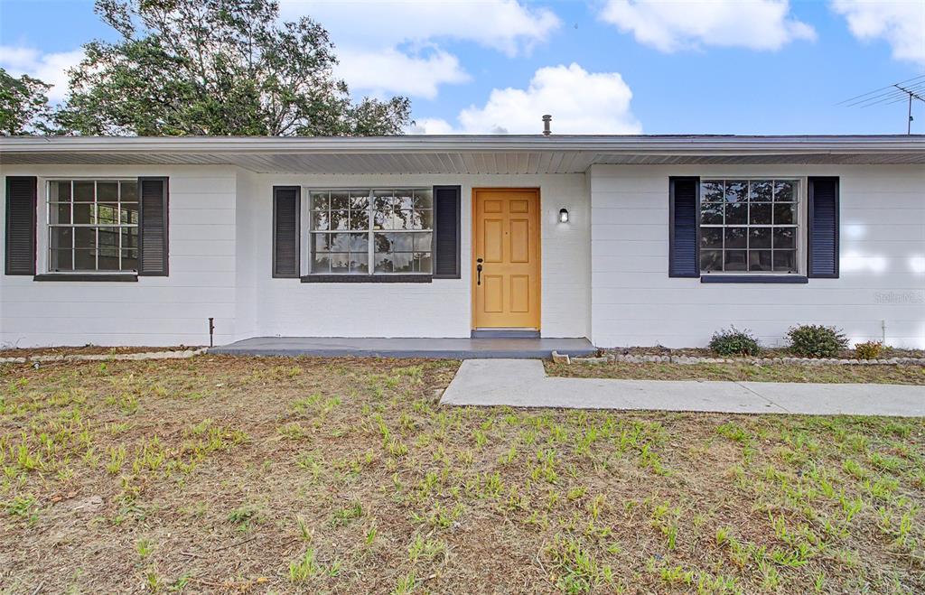 A single-story white house features a yellow front door, dark shutters, and a concrete walkway — 12368 MAYBERRY ROAD, SPRING HILL, FL