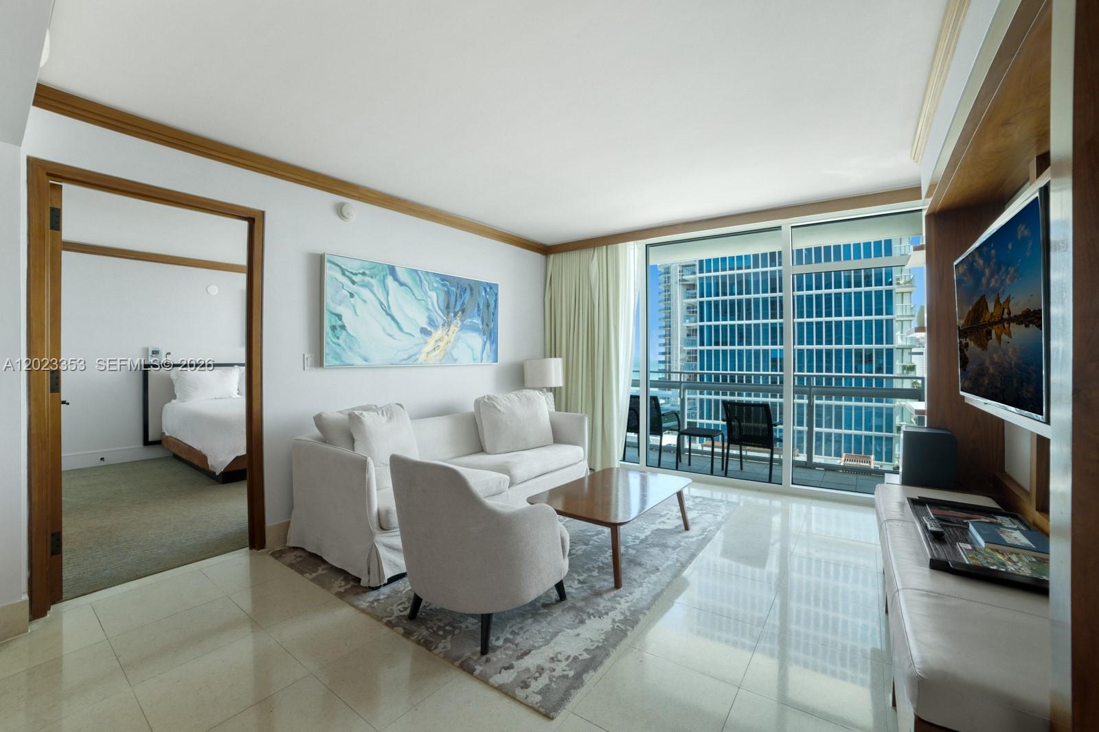 A living room with a white sofa, armchair, and balcony overlooking a modern glass building — 6801 Collins Ave, Miami Beach FL 33141, MIAMI BEACH, FL
