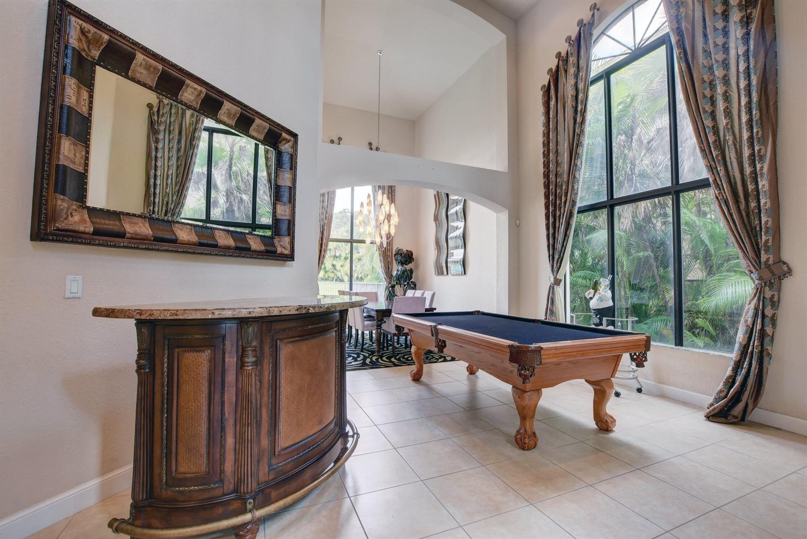 A room with a pool table, a curved wooden bar, and large windows with curtains — 3424 Collonade Drive, Wellington FL 33449, WELLINGTON, FL