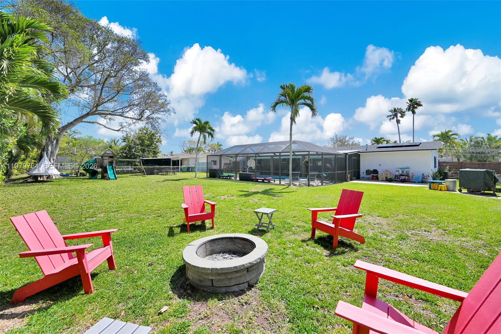 A grassy backyard features a stone fire pit, red Adirondack chairs, and a screened pool — 8935 SW 186th Ter, Cutler Bay FL 33157, CUTLER BAY, FL