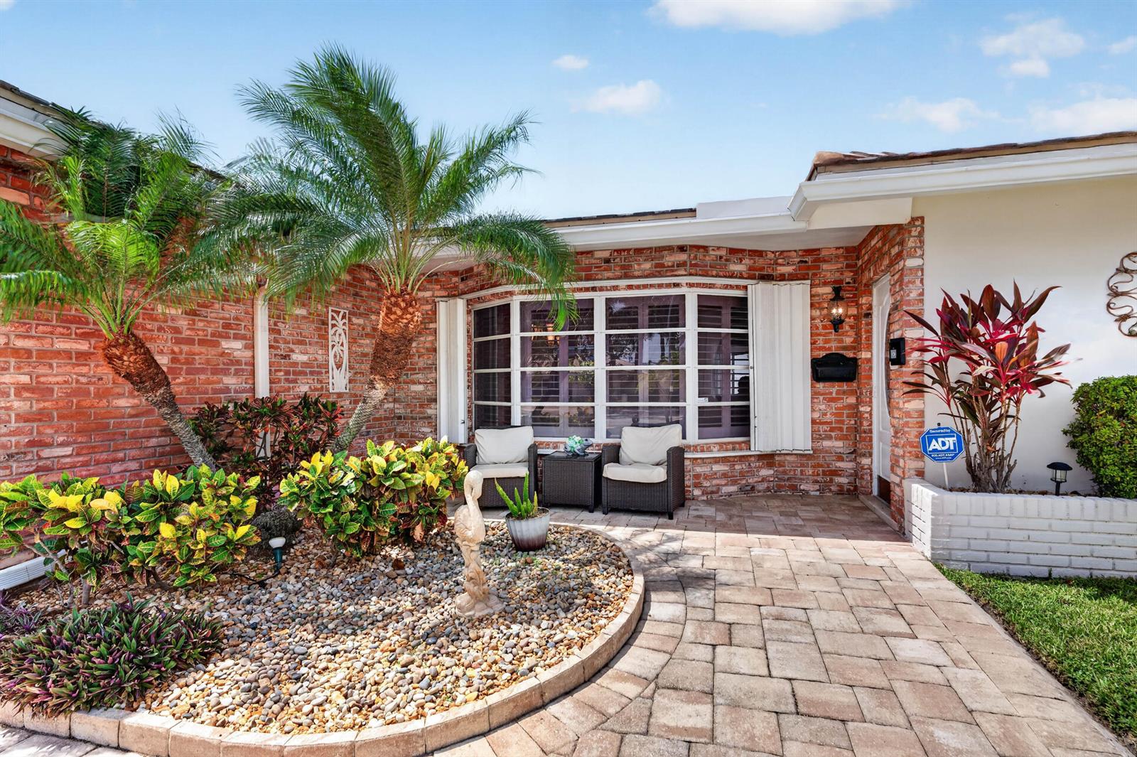 A brick house exterior features a paved walkway, a palm tree, and a small seating area — 1517 SE 14TH Street, Deerfield Beach FL 33441, DEERFIELD BEACH, FL