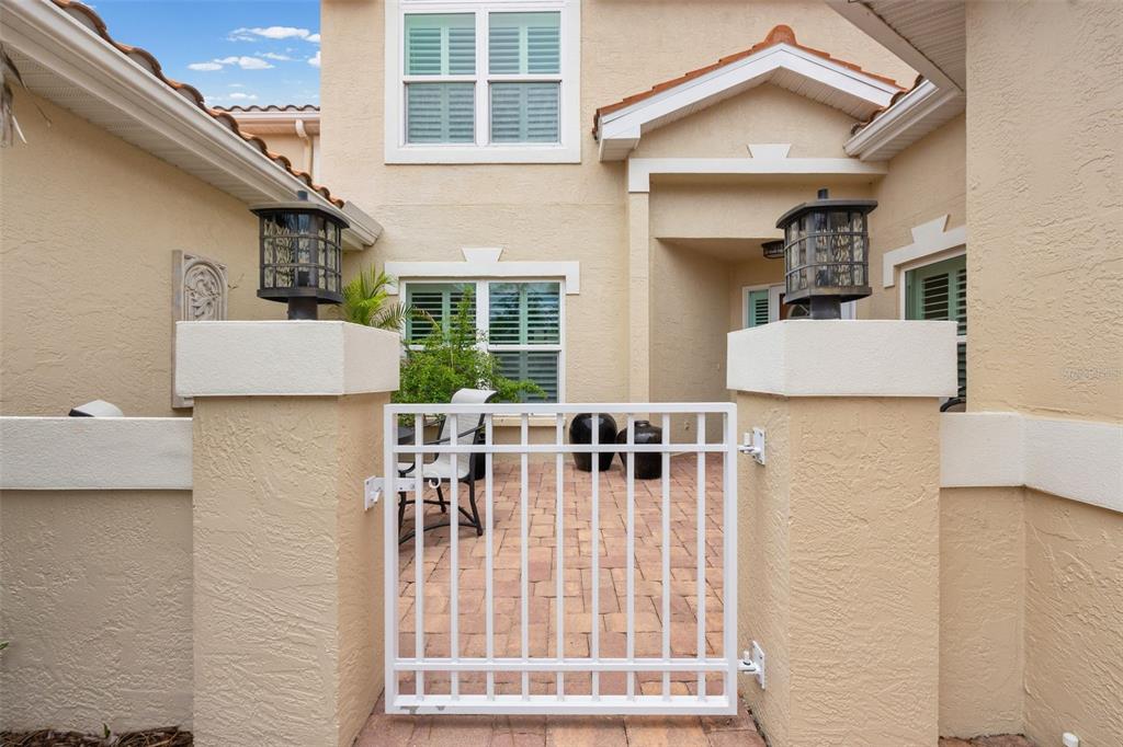 A white metal gate opens to a paved courtyard in front of a tan stucco house — 264 WOODS POINT ROAD, OSPREY, FL