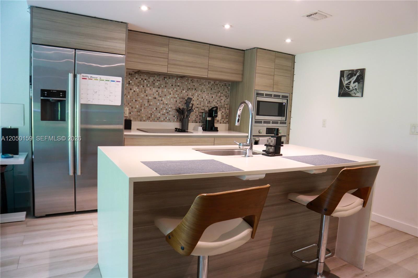 A modern kitchen features a white island with two stools, stainless steel appliances, and wood-toned cabinetry — 6620 Indian Creek Dr # 302, Miami Beach FL 33141, MIAMI BEACH, FL