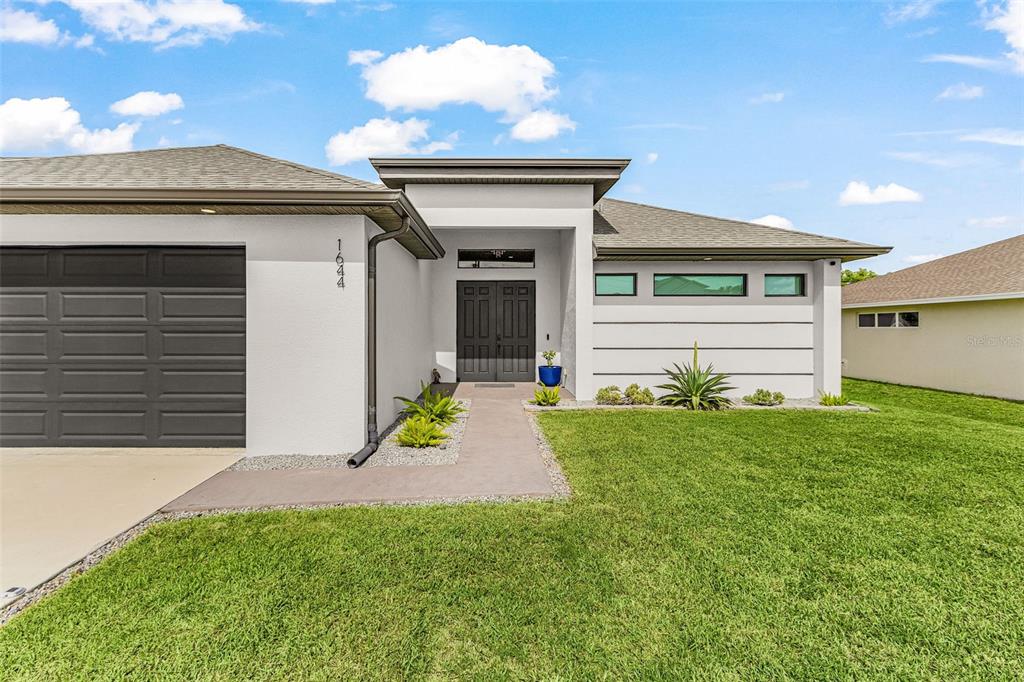 A modern single-story house exterior features a gray garage, dark front door, and horizontal windows — 1644 NW 37TH AVENUE, CAPE CORAL, FL