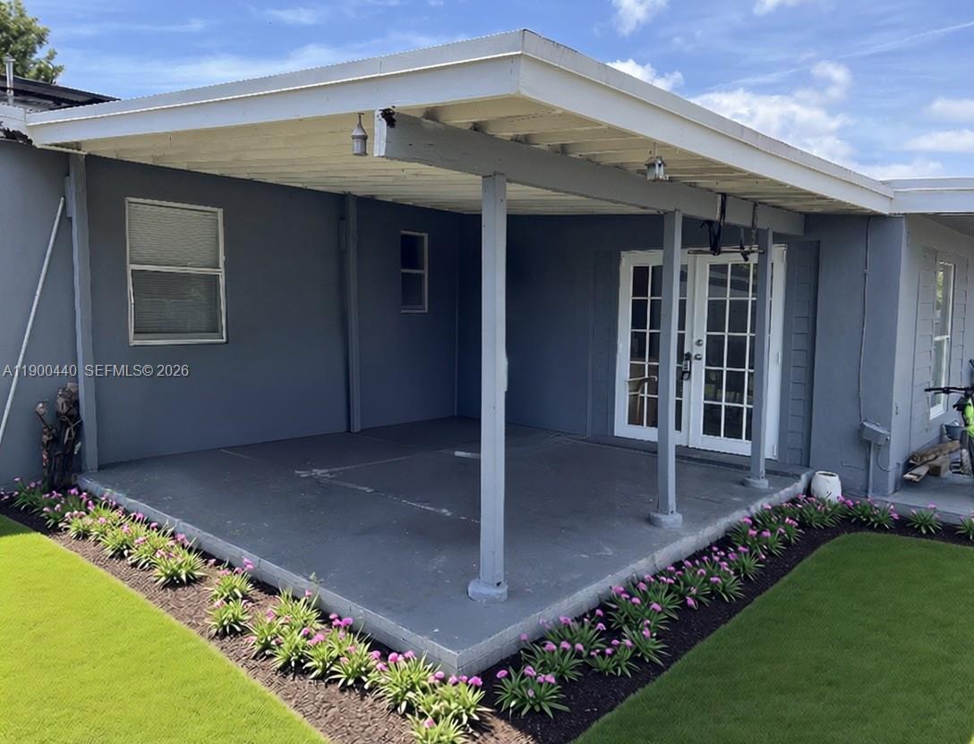 A covered concrete patio features a gray exterior wall, white French doors, and landscaped flowerbeds — 1885 NE 53rd St, Pompano Beach FL 33064, POMPANO BEACH, FL