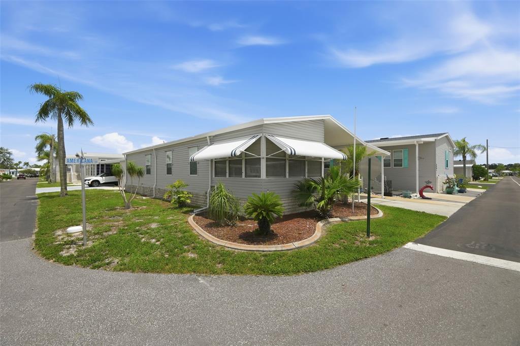 A light gray manufactured home with a white awning stands on a corner lot — 6907 AMERICANA DRIVE NE UNIT 101, ST PETERSBURG, FL