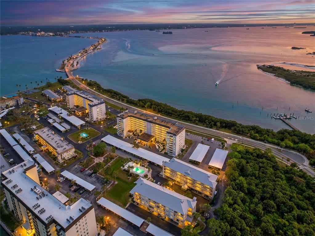 An aerial view shows multiple apartment buildings, swimming pools, and a causeway over the water — 7 ISLAND PARK PLACE UNIT 204, DUNEDIN, FL
