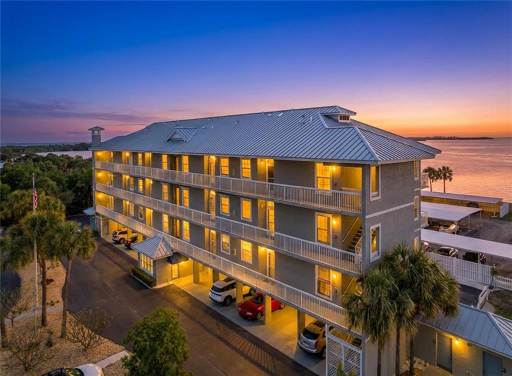 An elevated multi-story apartment building with covered parking sits near the water at sunset — 7 ISLAND PARK PLACE UNIT 204, DUNEDIN, FL