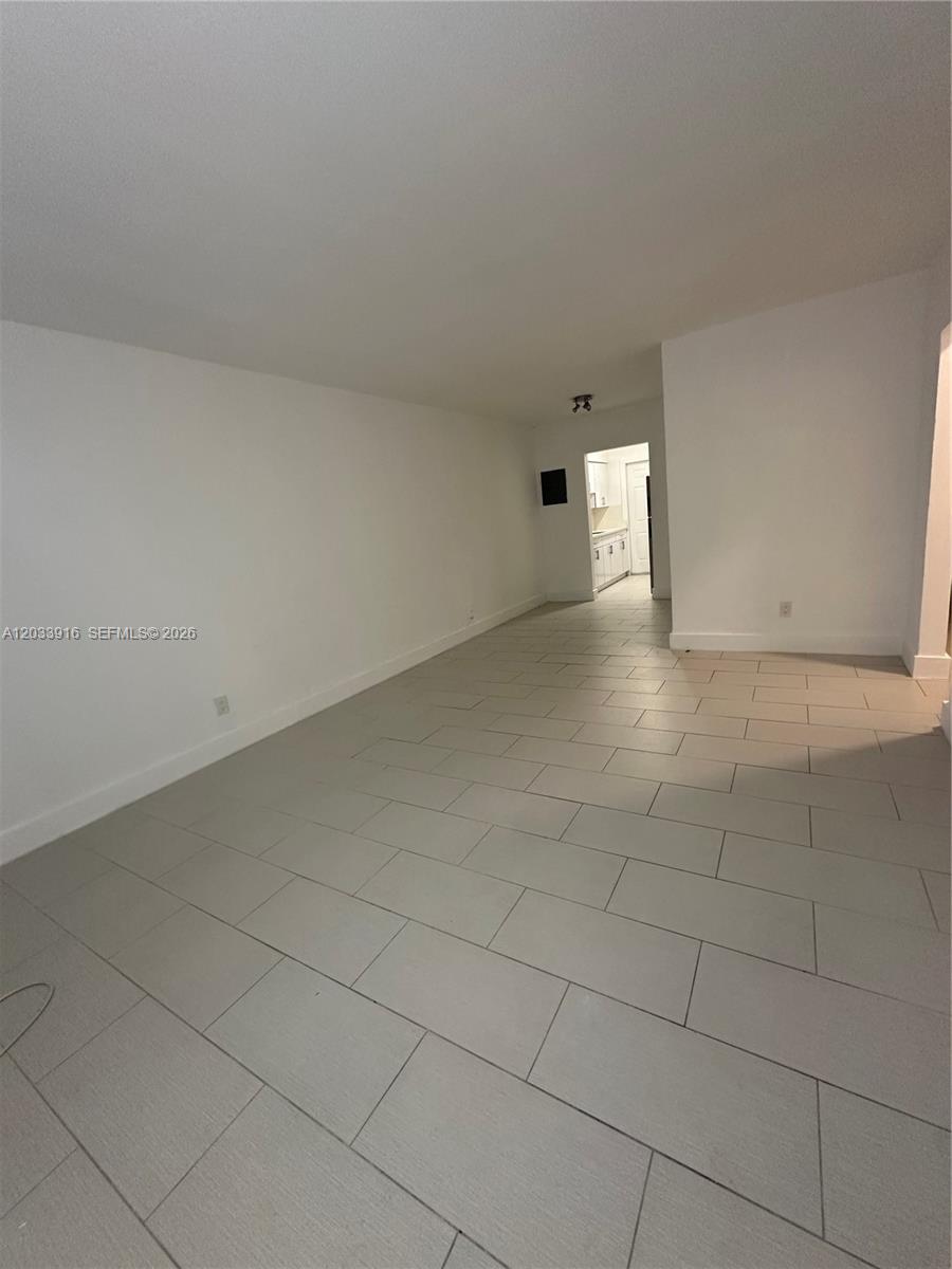 An empty room features light gray tile flooring, white walls, and a doorway to a kitchen — 8205 Crespi Blvd # 3, Miami Beach FL 33141, MIAMI BEACH, FL