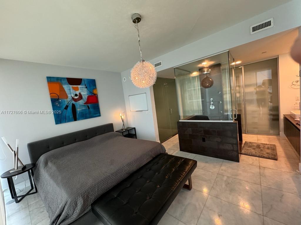 A bedroom features a bed, a black bench, modern lighting, and an open glass-enclosed bathroom — 1040 Biscayne Blvd # 1505, Miami FL 33132, MIAMI, FL