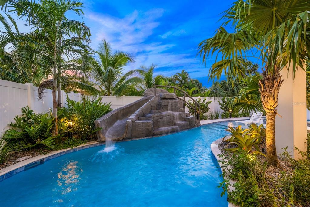 A swimming pool features a rock-style slide and a bridge surrounded by lush tropical landscaping — 6910 MIDNIGHT PASS ROAD UNIT A, SARASOTA, FL