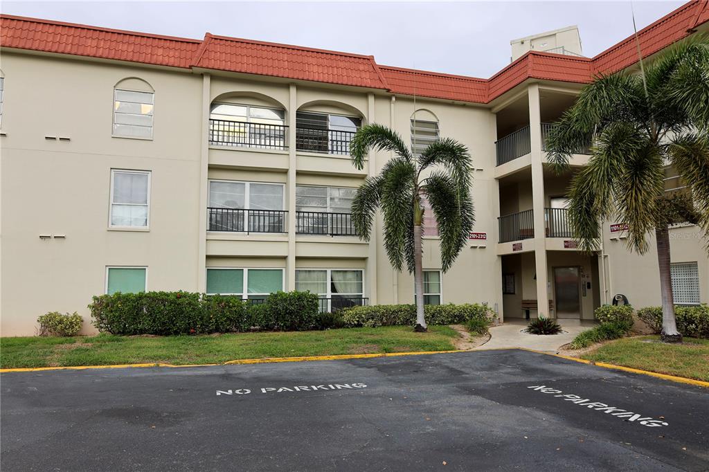 A three-story apartment building with a red tile roof and a paved parking area — 2700 BAYSHORE BOULEVARD UNIT 2105, DUNEDIN, FL