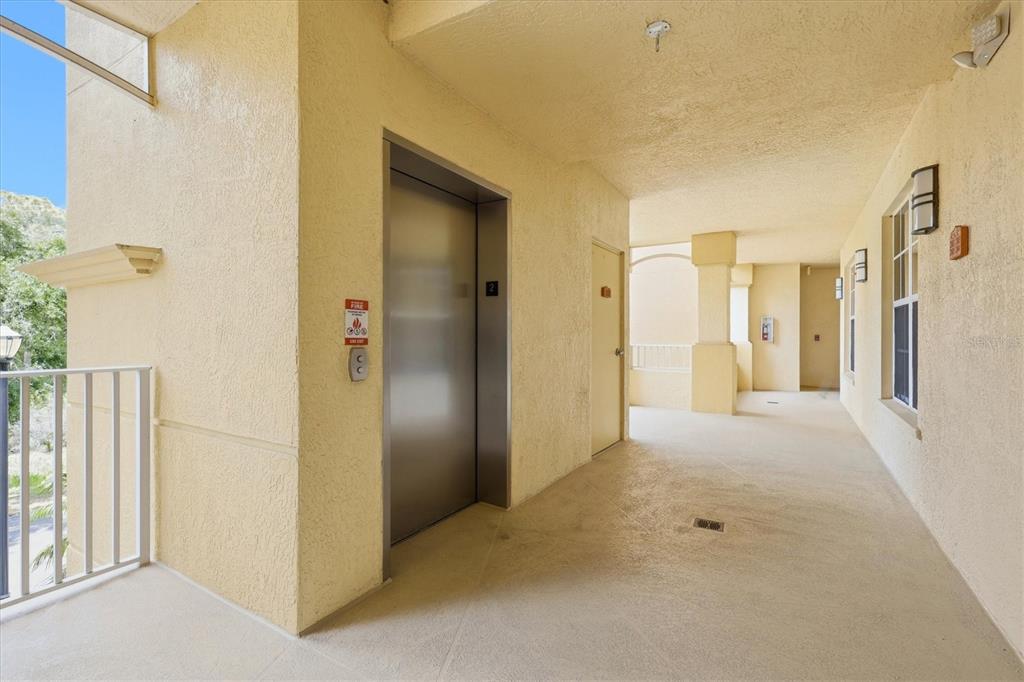 An outdoor apartment building hallway features a stainless steel elevator, beige walls, and tile flooring — 6360 WATERCREST WAY UNIT 201, LAKEWOOD RANCH, FL