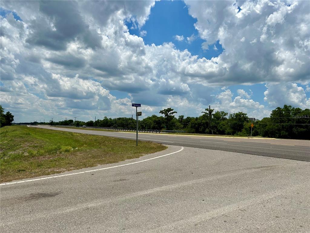 A paved road intersection features a street sign and a grassy shoulder under a cloudy sky — BRONCO DRIVE, ZOLFO SPRINGS, FL