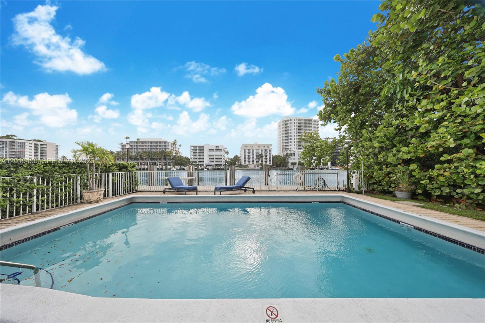 A rectangular swimming pool sits on a patio overlooking a waterway and distant apartment buildings — 34 Bal Bay Dr, Bal Harbour FL 33154, BAL HARBOUR, FL