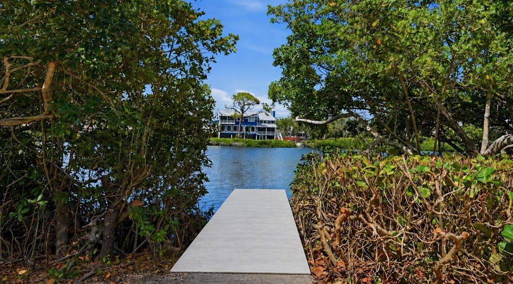 A wooden dock extends into a calm waterway toward a blue house framed by trees — 7723 SANDERLING ROAD, SARASOTA, FL