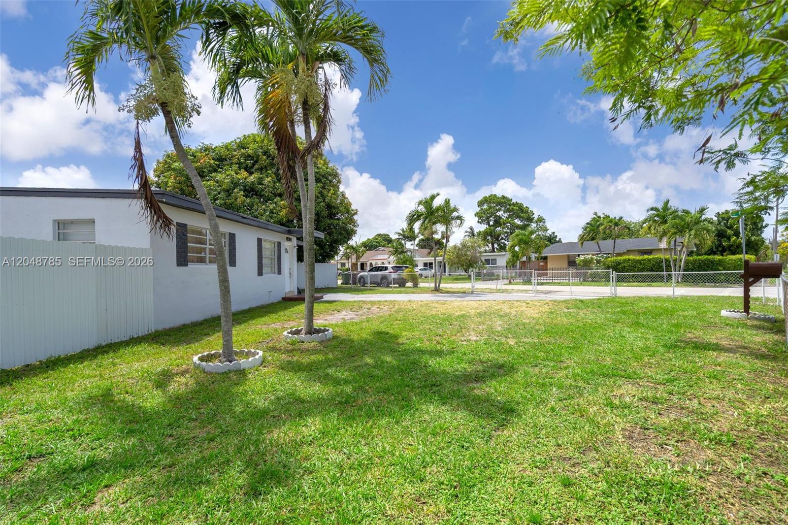 A grassy yard features two palm trees beside a white house with a fence — 6084 E 7th Ave, Hialeah FL 33013, HIALEAH, FL
