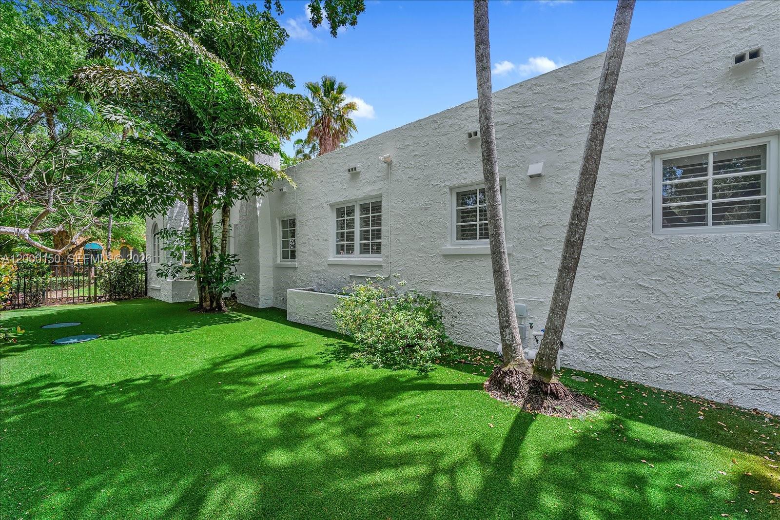 The exterior of a white stucco house features a green artificial turf lawn and trees — 1102 Genoa St, Coral Gables FL 33134, CORAL GABLES, FL