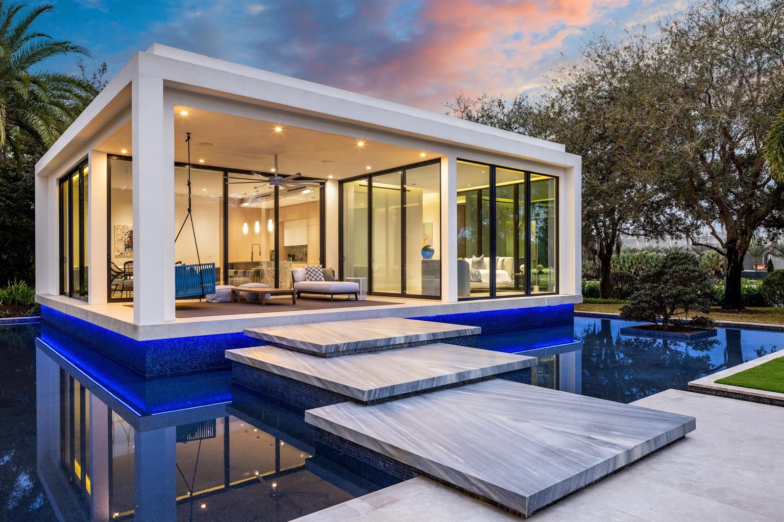 A modern glass-walled pavilion sits on a pool with stone stepping stones at sunset — 16191 Quiet Vista Circle, Delray Beach FL 33446, DELRAY BEACH, FL