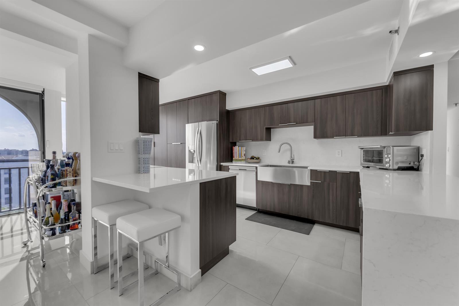 A modern kitchen features dark wood cabinets, white countertops, a farmhouse sink, and bar seating — 7178 Promenade Drive # 801b, Boca Raton FL 33433, BOCA RATON, FL