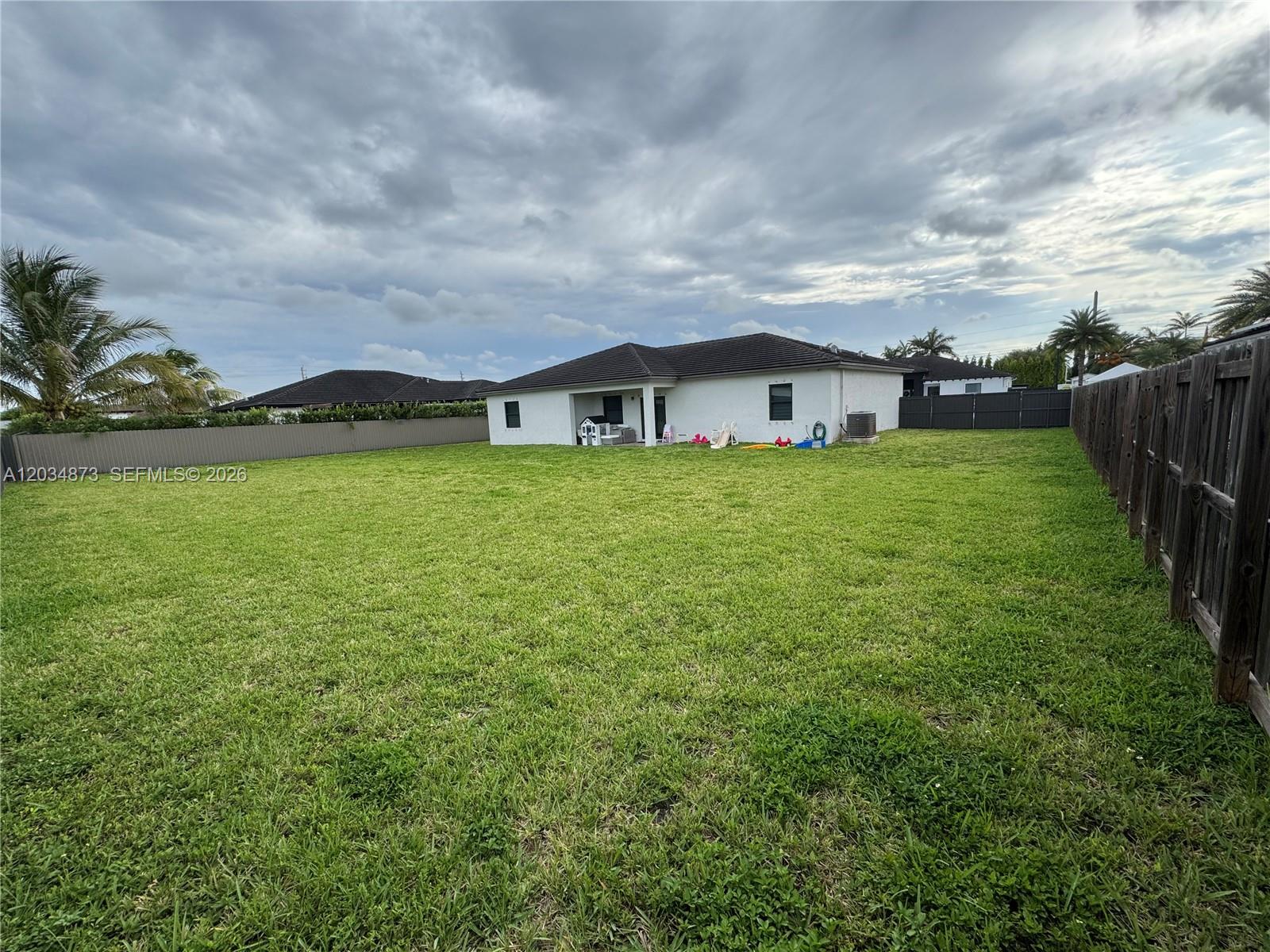 A large, grassy backyard features a white house with a covered patio and wooden fencing — 21483 SW 132nd Pl, Miami FL 33177, MIAMI, FL