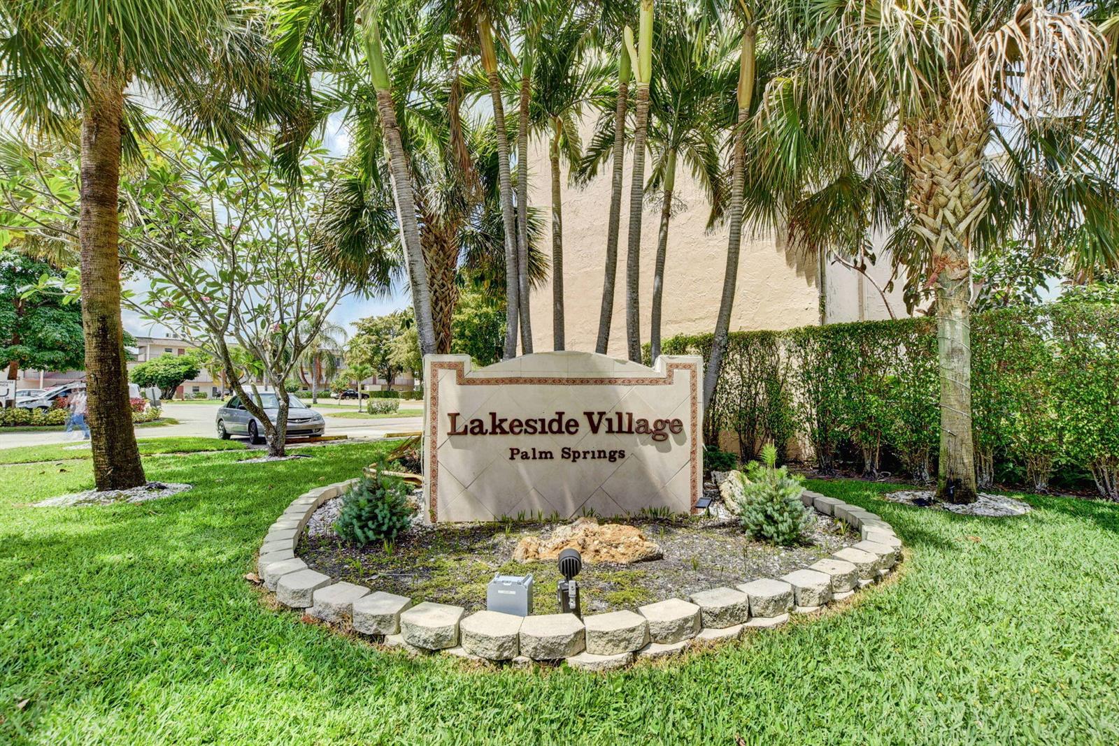 A stone sign for Lakeside Village Palm Springs sits in a landscaped garden with palm trees — 725 Lori Drive # 106, Palm Springs FL 33461, PALM SPRINGS, FL