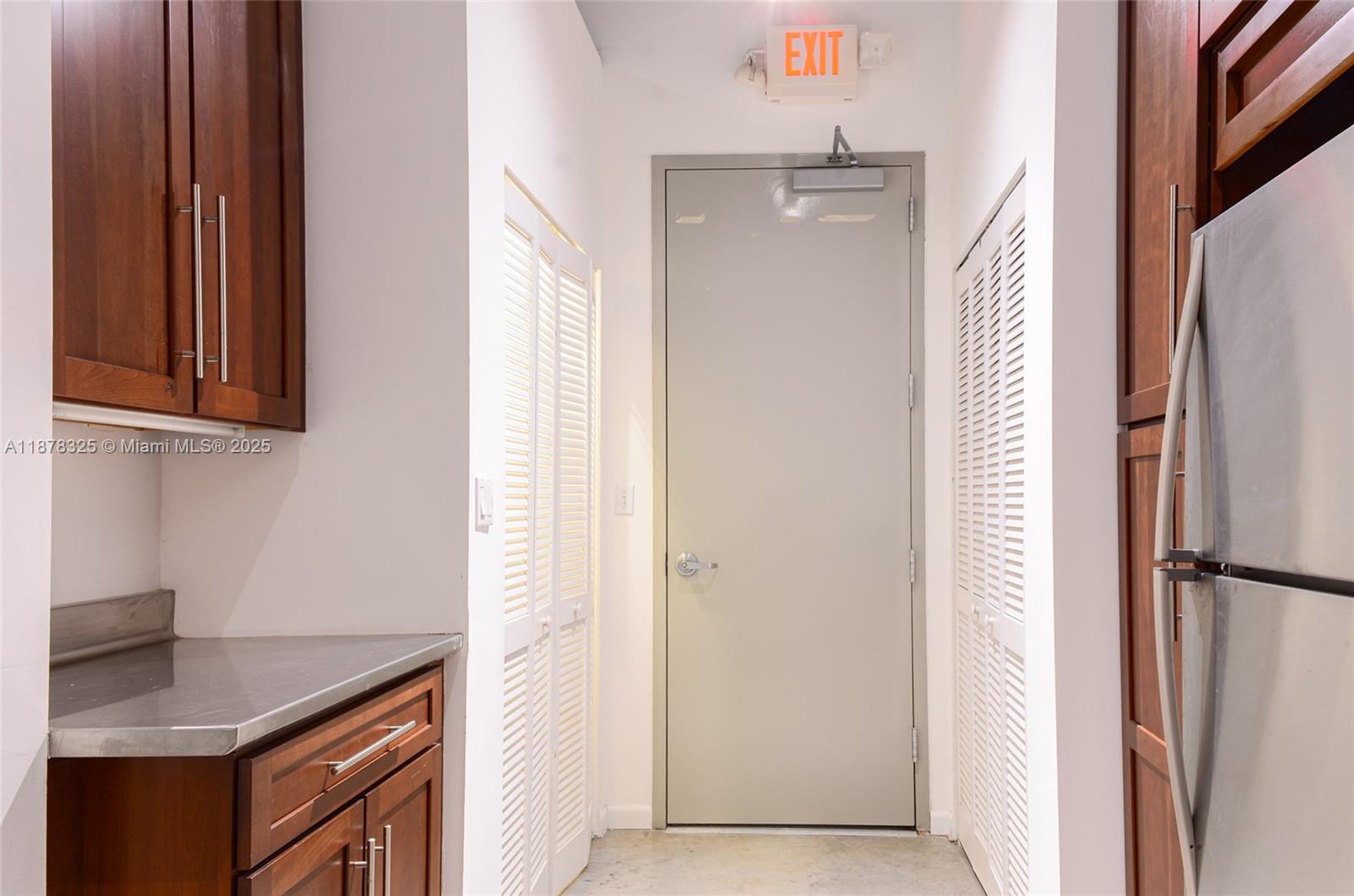 A kitchen area features wooden cabinets, a stainless steel refrigerator, and a gray exit door — 250 NW 23rd st # 308, Miami FL 33127, MIAMI, FL