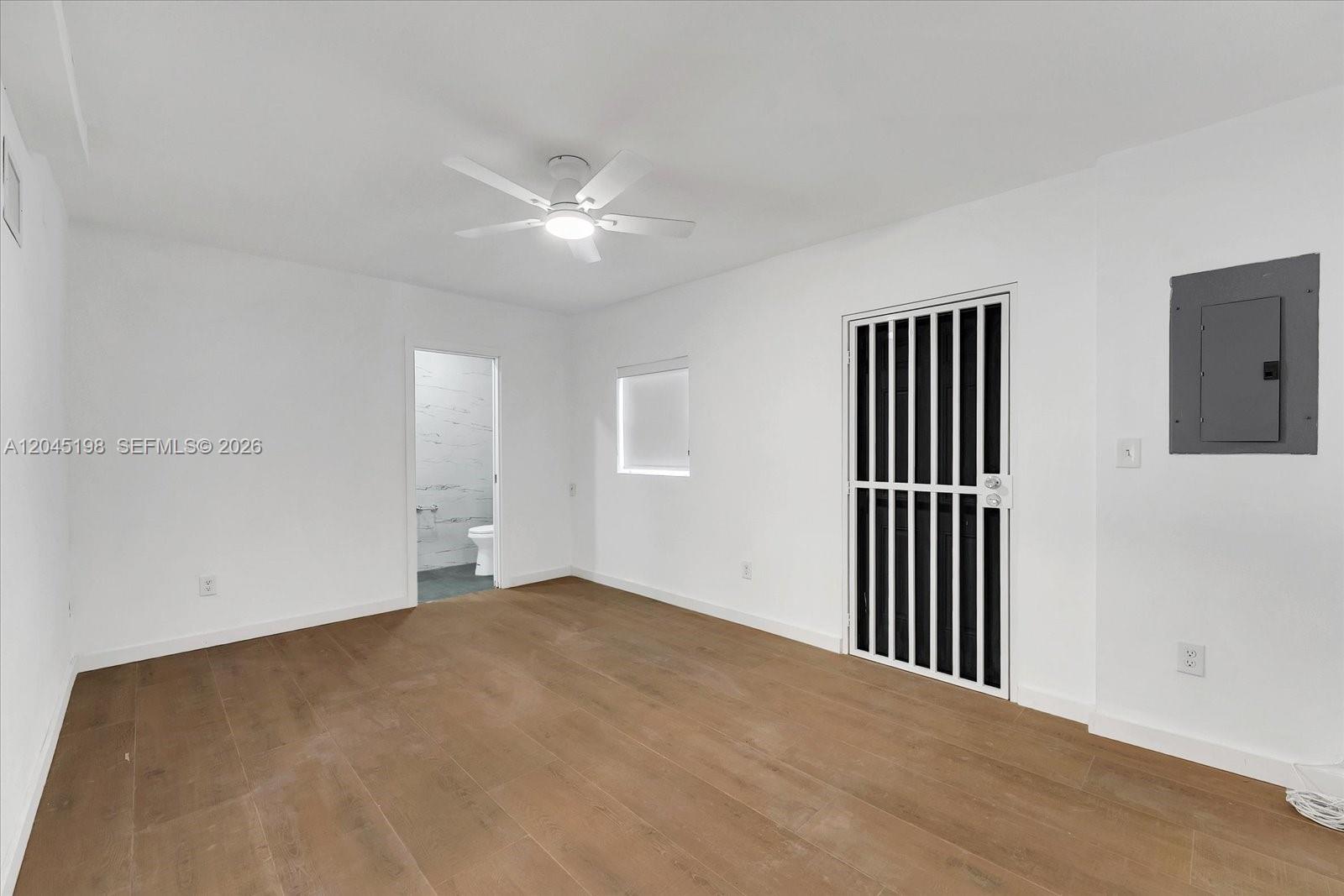 A white-walled room features wood-look flooring, a ceiling fan, a bathroom doorway, and a security door — 13415 NW 22nd Ave, Miami FL 33167, MIAMI, FL