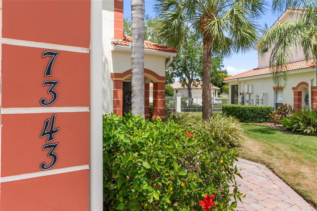 A vertical house number sign is mounted on a reddish-orange exterior wall near a walkway — 7343 FOUNTAIN PALM CIRCLE UNIT na, BRADENTON, FL