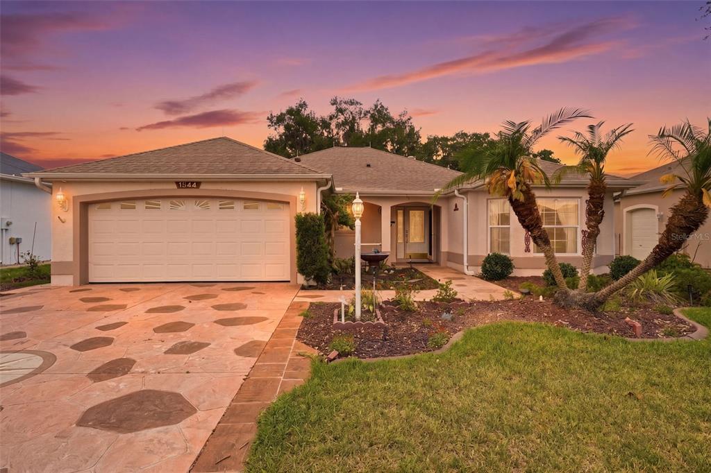 A single-story house with a two-car garage, a paved driveway, and palm trees at sunset — 1344 AUGUSTINE DRIVE, THE VILLAGES, FL