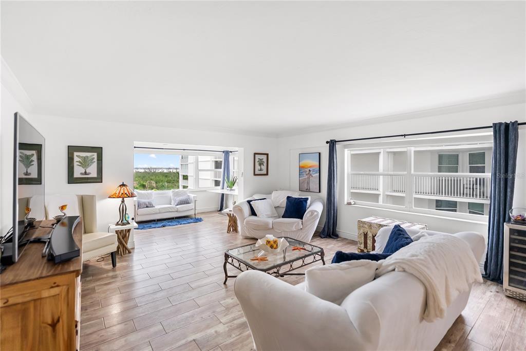 A bright living room features white sofas, wood-look tile flooring, and large windows with views — 1750 BENJAMIN FRANKLIN DRIVE UNIT 6G, SARASOTA, FL