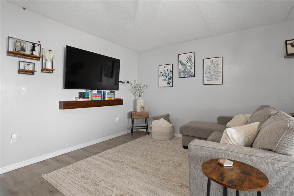 A living room features a wall-mounted television, a floating wooden shelf, and a gray sectional — 1154 PASEO DEL SOL UNIT B, CASSELBERRY, FL