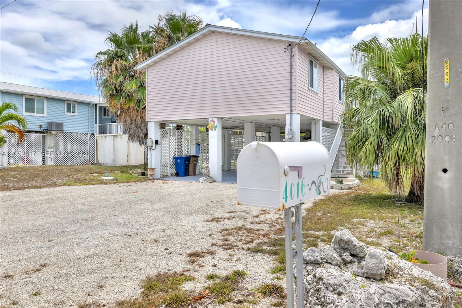 A stilt house with light pink siding stands behind a mailbox on a gravel lot — LOWER KEYS, FL