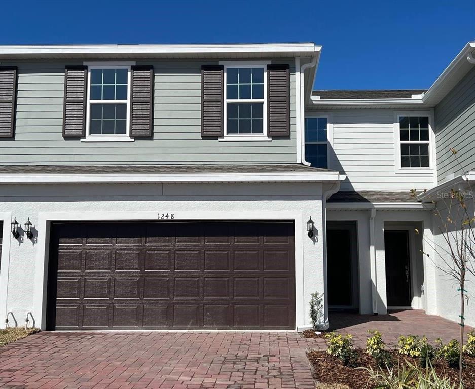 A two-story townhouse exterior features a brown garage door, light siding, and a brick driveway — 1248 WISPY CYPRESS DRIVE, KISSIMMEE, FL