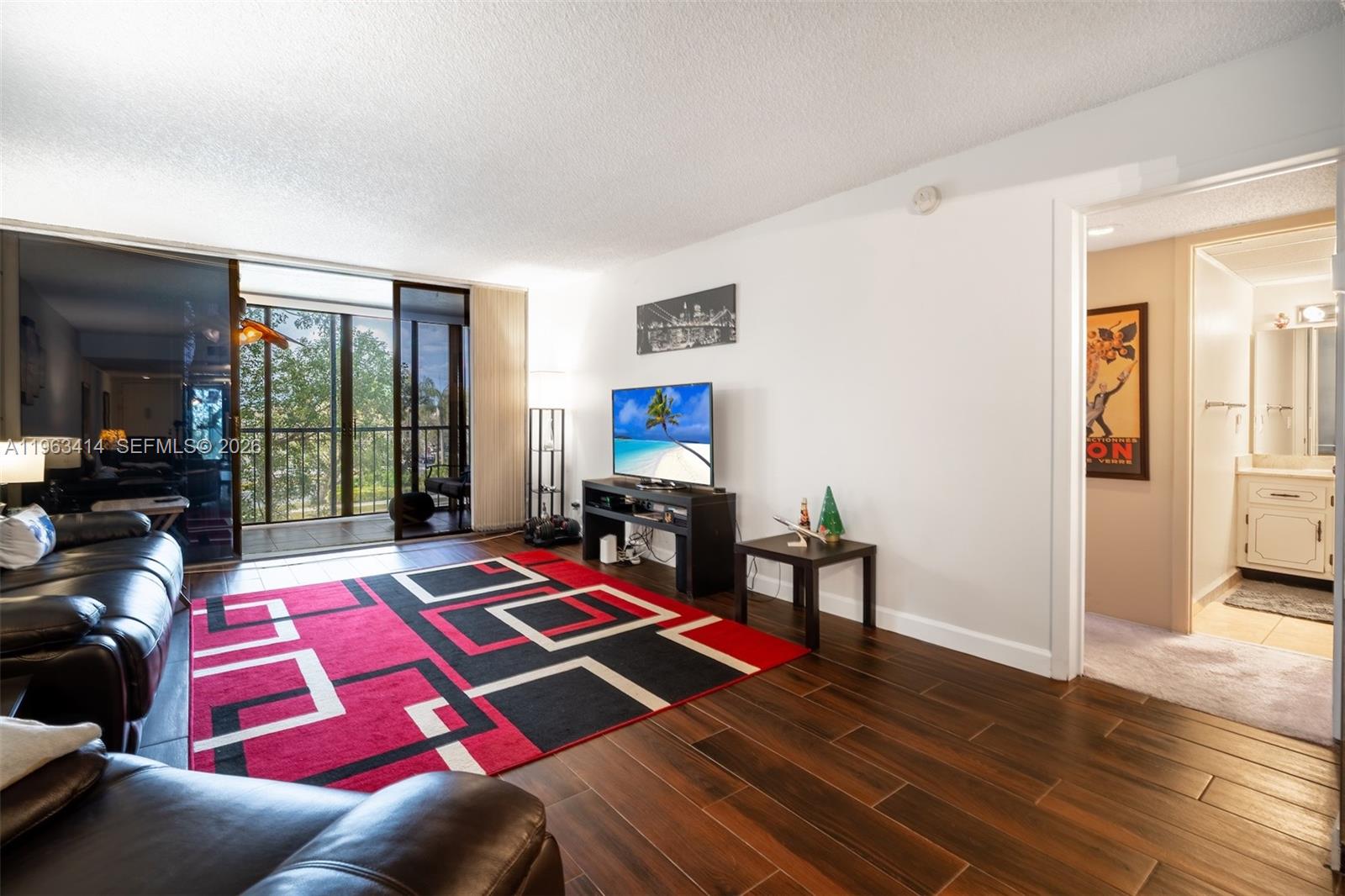 A living room features dark wood-look tile flooring, a red geometric rug, and a balcony — 16475 Golf Club Rd # 307, Weston FL 33326, WESTON, FL