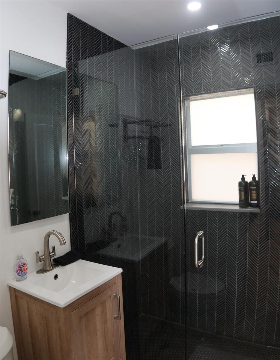 A bathroom features a wood vanity, a mirror, and a shower with dark herringbone tiles — 3408 Spring Street # 2, Pompano Beach FL 33062, POMPANO BEACH, FL