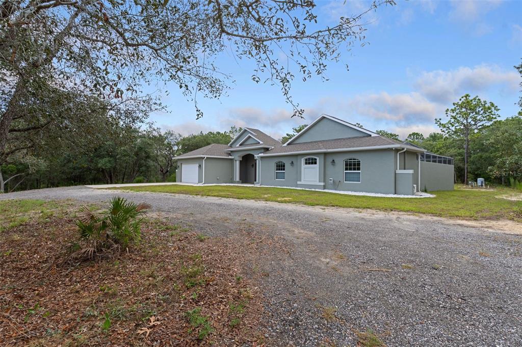 A single-story gray house with a gravel driveway and a garage in a wooded area — 14610 HIDDEN DEER TRAIL, BROOKSVILLE, FL