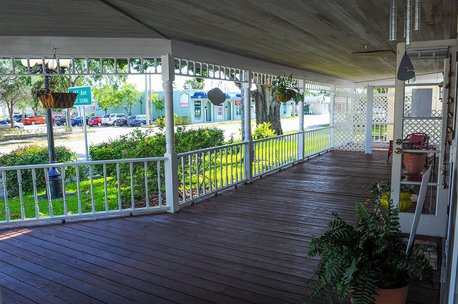 A wide, covered wooden porch features white railings, hanging plants, and a view of street traffic — 1443 19th Place, Vero Beach FL 32960, VERO BEACH, FL
