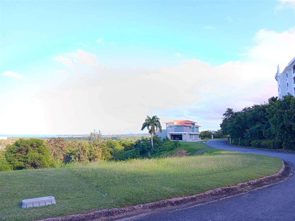 A paved road curves past a grassy lot toward a modern house with ocean views — 6000 RIO MAR BOULEVARD Lot 53, RIO GRANDE, PR