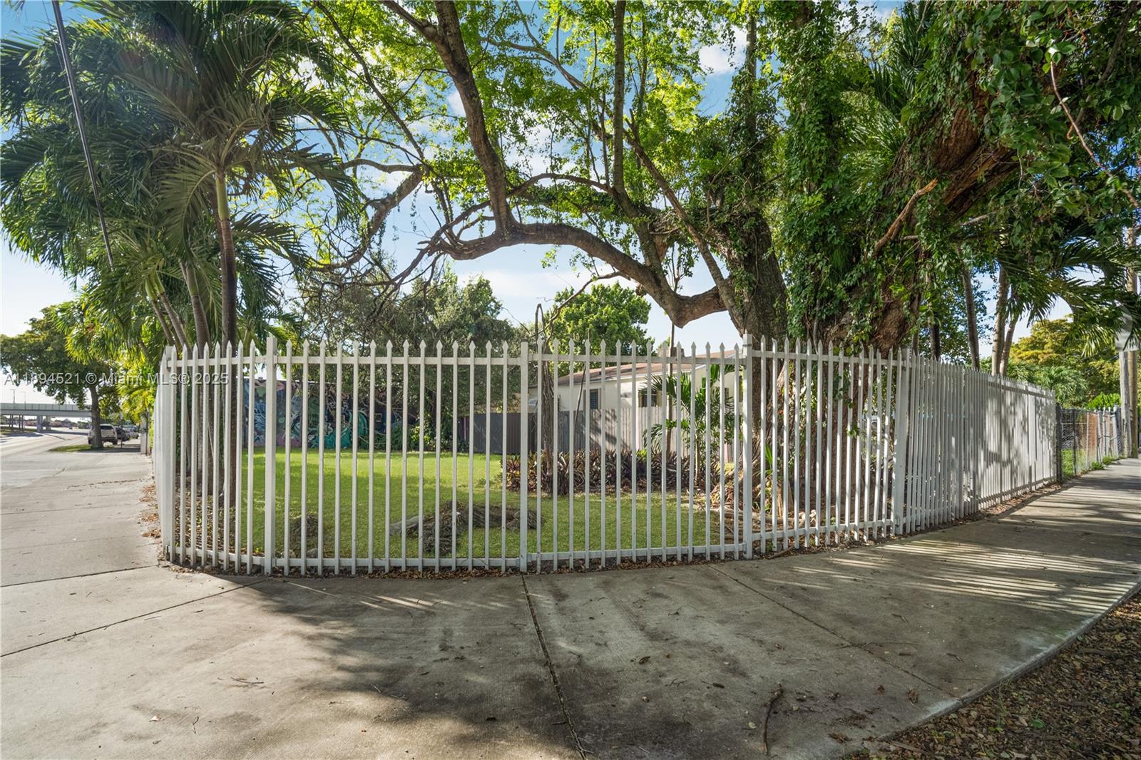 A white metal fence encloses a grassy corner lot with large trees and a house — 1121 NW 29th St, Miami FL 33127, MIAMI, FL