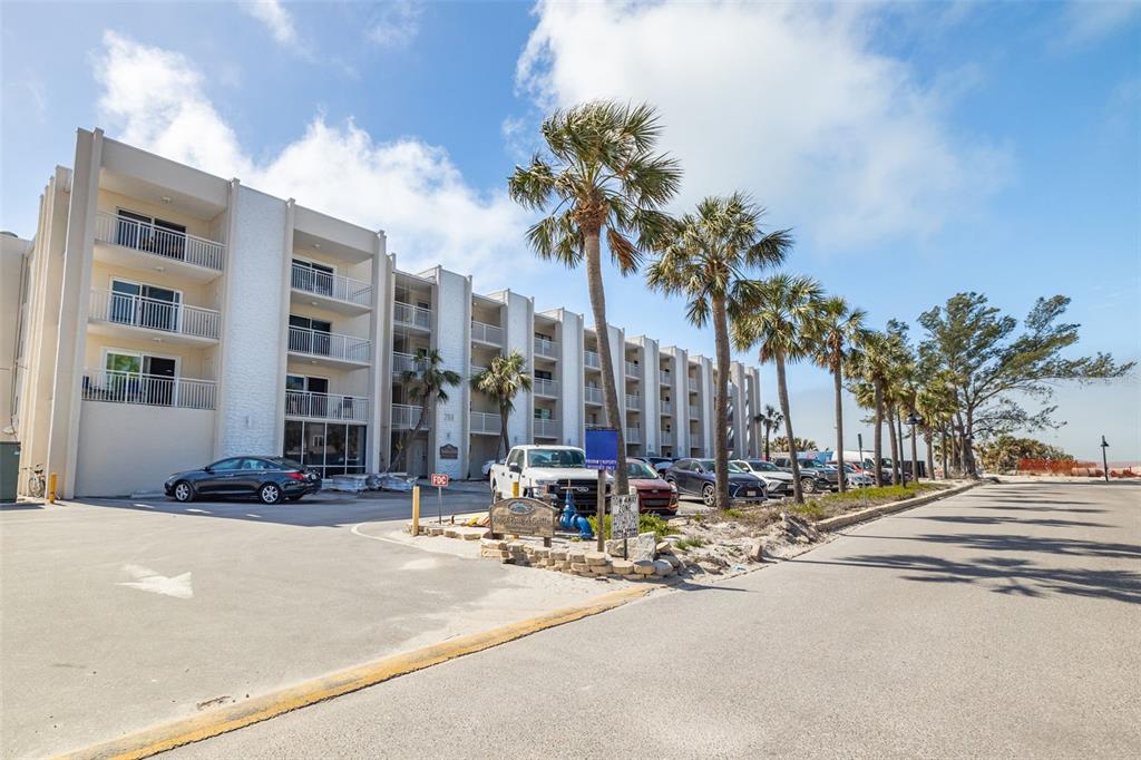 A multi-story white apartment building with balconies stands next to a parking lot and palms — 200 1ST AVENUE UNIT 212, ST PETE BEACH, FL