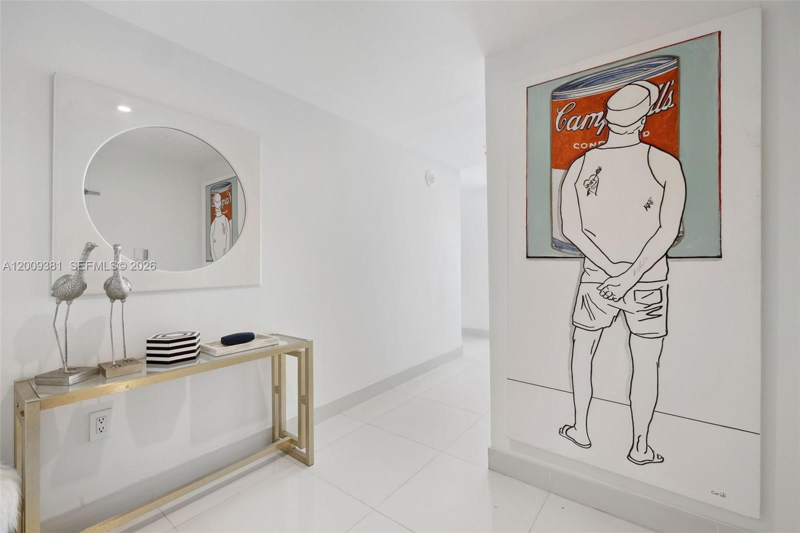 A white hallway features a gold console table, a round mirror, and large pop-art painting — 465 Brickell Ave # 701, Miami FL 33131, MIAMI, FL