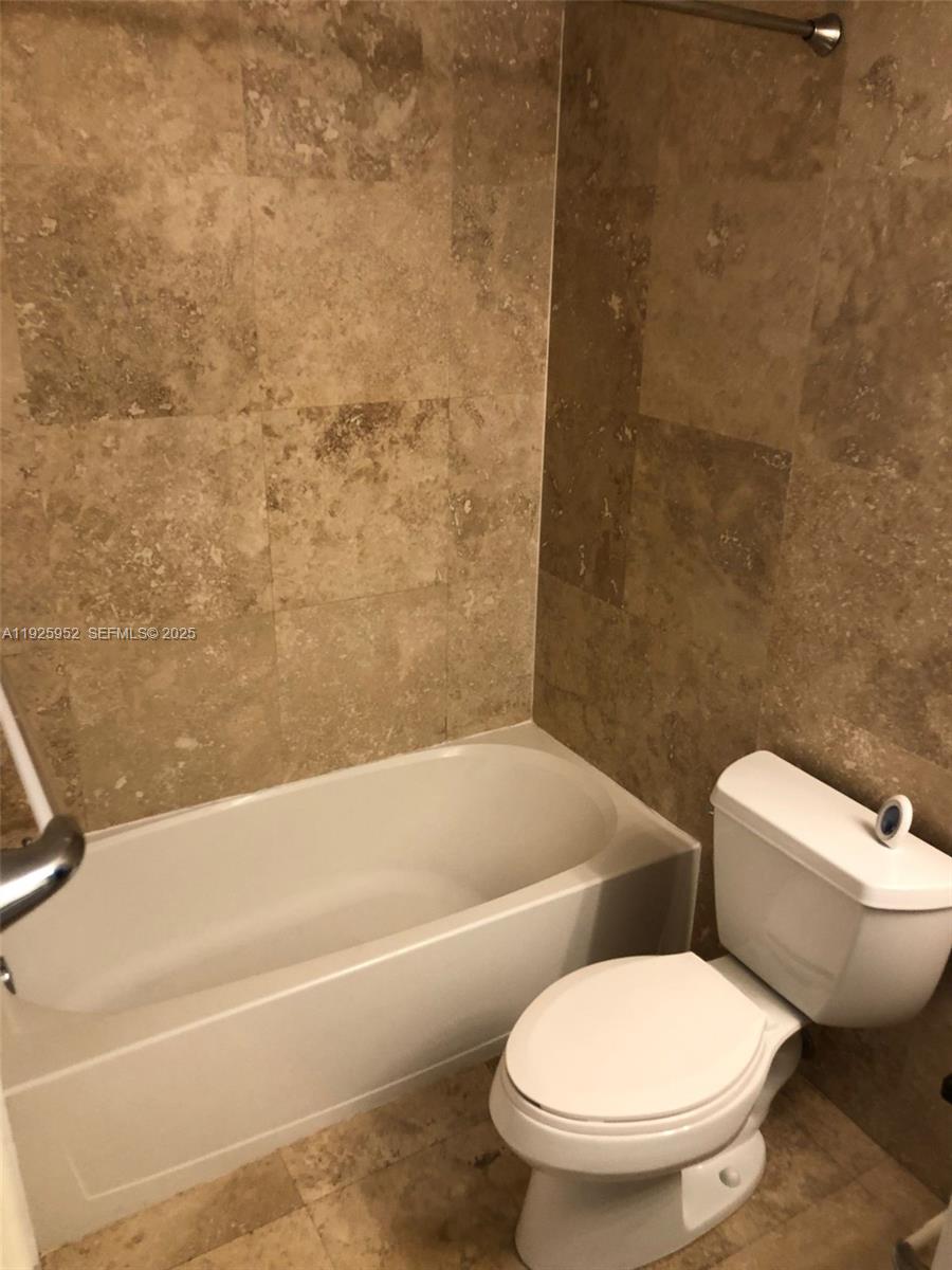 A bathroom features a white bathtub, a white toilet, and tan stone wall tiles — 1650 Brickell Ave # 111, Miami FL 33129, MIAMI, FL