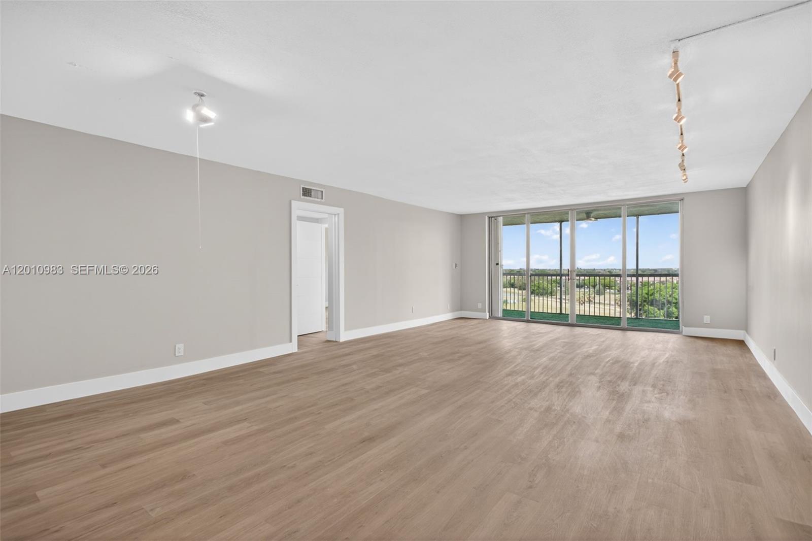 An empty living room features light wood-look flooring, neutral walls, and a sliding glass door — 3090 N Course Dr # 807, Pompano Beach FL 33069, POMPANO BEACH, FL